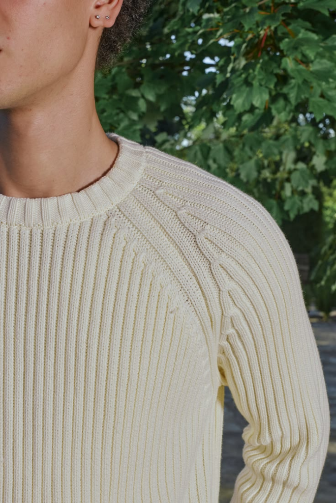 Close-up of a cream ribbed knit sweater on the shoulder and neckline, with greenery in the background.