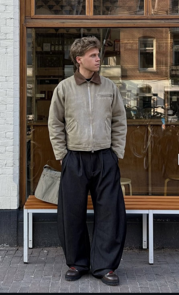 Man in a tan canvas jacket and black wide-leg trousers standing outside a storefront, holding a tan bag.