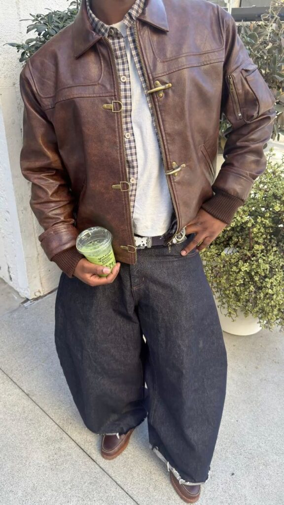 Man wearing a brown leather trucker jacket over a cream henley with wide-leg dark denim jeans and brown boots, leaning against a wall.