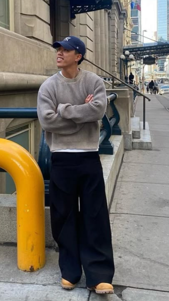Man in a gray ribbed crewneck sweater, navy cap, and black wide-leg trousers standing on a city sidewalk.